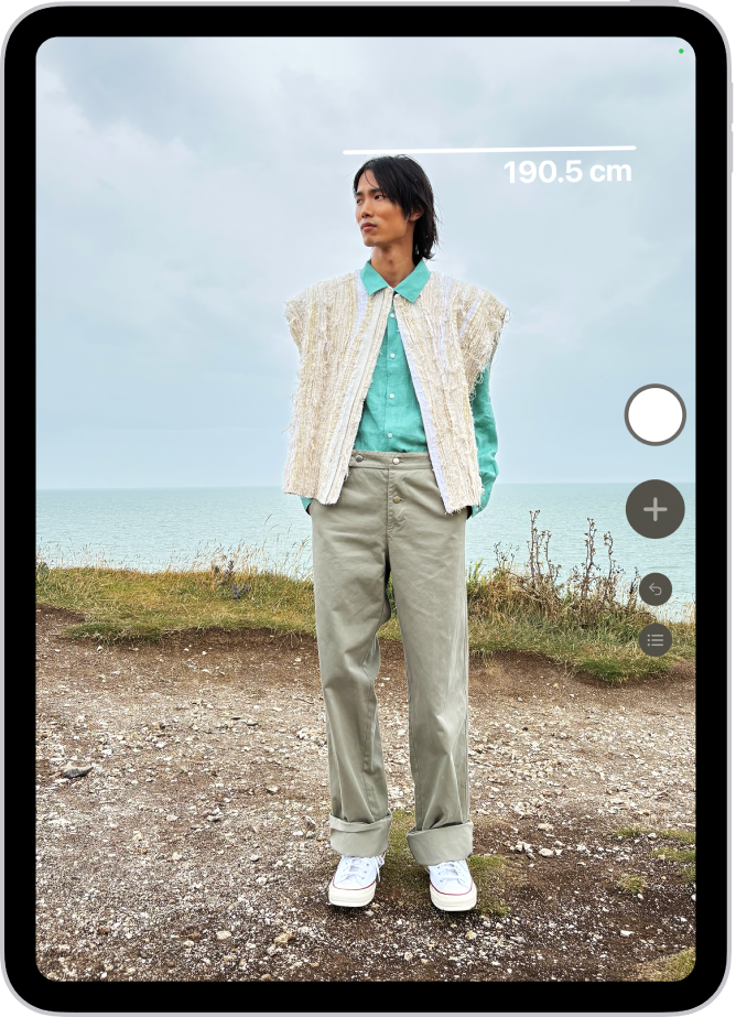 Chiều cao của một người đang được đo trong ứng dụng Đo, với số đo chiều cao đang hiển thị ở trên đỉnh đầu của người đó. Nút Chụp ảnh ở gần góc phía dưới bên phải để chụp ảnh phép đo. Chỉ báo Camera đang được sử dụng màu lục xuất hiện ở trên cùng.