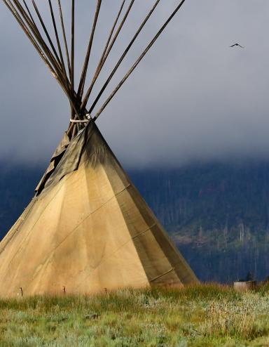 A large tipi sits in a field of grass. Its poles rise into a misty sky. Partially obscured.