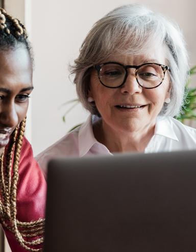 A young woman who looks to be in her teen years smiles as she works at a computer alongside an older, white-haired woman who is also smiling. Partially obscured.