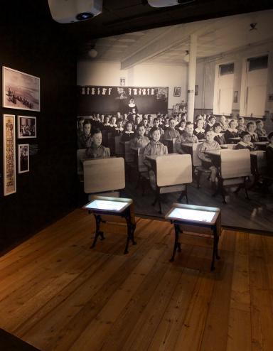 A Museum exhibit showing a black-and-white photo of children sitting in rows at school desks. Two desks, similar to those in the photo sit in the centre of the exhibit. A headline on a text panel reads “Childhood Denied.” Partially obscured.