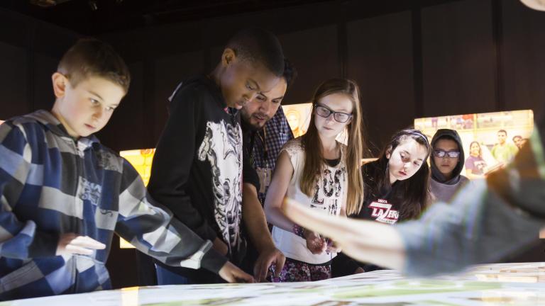 A group of students look at the top of an interactive table emitting white light. Partially obscured.