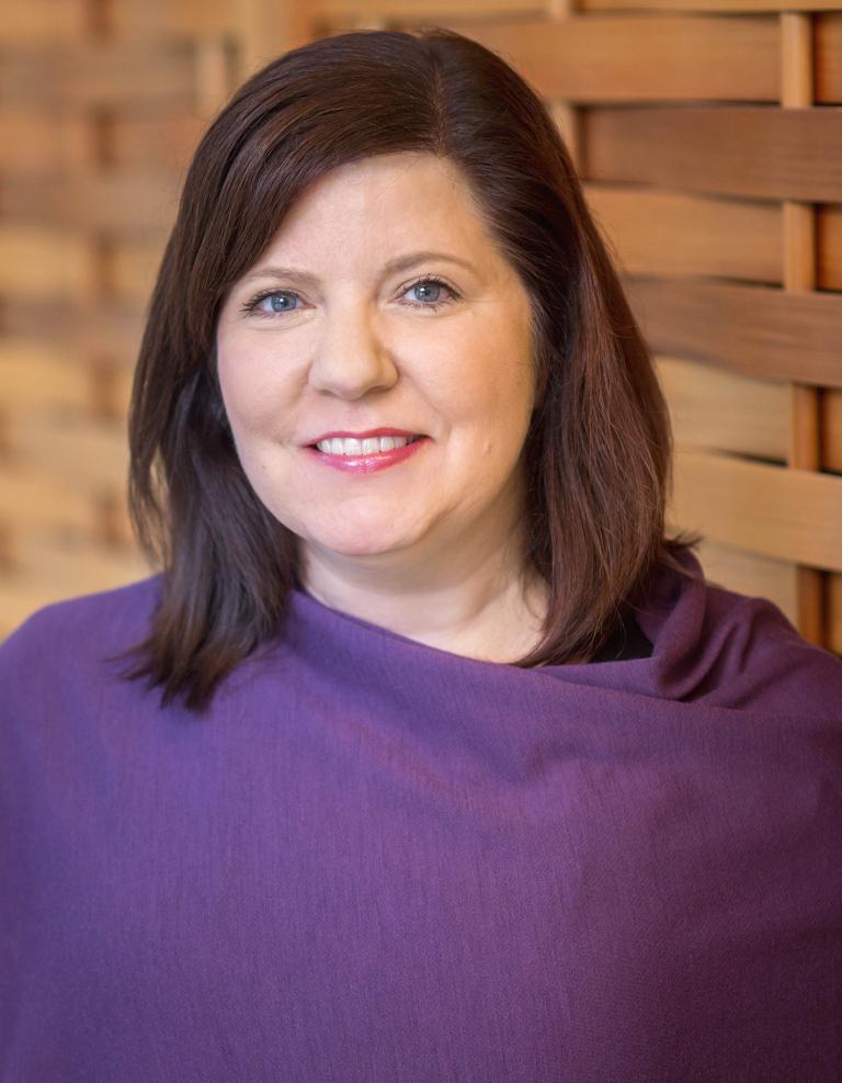 A woman with dark hair and blue eyes smiles warmly at the camera. She's wearing a purple shawl, and is positioned in front of a wooden lattice wall. Partially obscured.