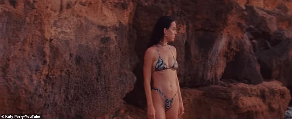 As the sunsets, a bikini-clad Perry flashes her backside as she climbs on red rocks on the shore to then cannonball back into the water