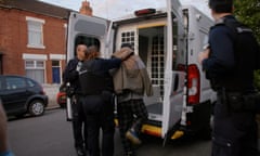 A migrant being put into the back of a white security van while being held by an immigration officer