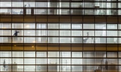 Close-up of a modern office building at night with  a figure on the phone silhouetted against one of the illuminated windows