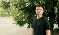 A Pakistani man standing in a slightly overgrown car park in London with a tree behind him