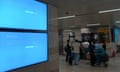 Two large screens in a polished granite wall, both blank, with people carrying suitcases beyond it.