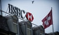 HSBC sign and red and white logo, plus a red and white Swiss flag, on the roof of one of the bank's premises in Geneva. They are seen against a pale, grey sky in which a bird hovers