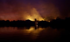 Fire illuminates smoke rising above a nighttime horizon of trees on a lake