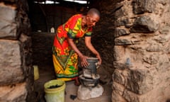 An older woman bends over a small stove using solid fuel