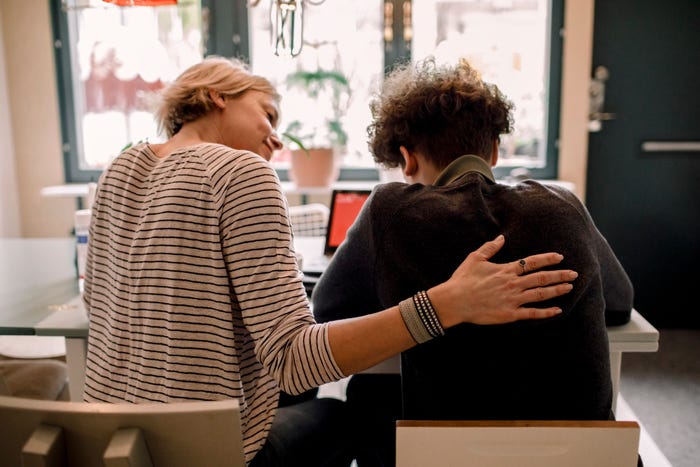 Woman with arm around teenage son, sitting at table, view from behind.