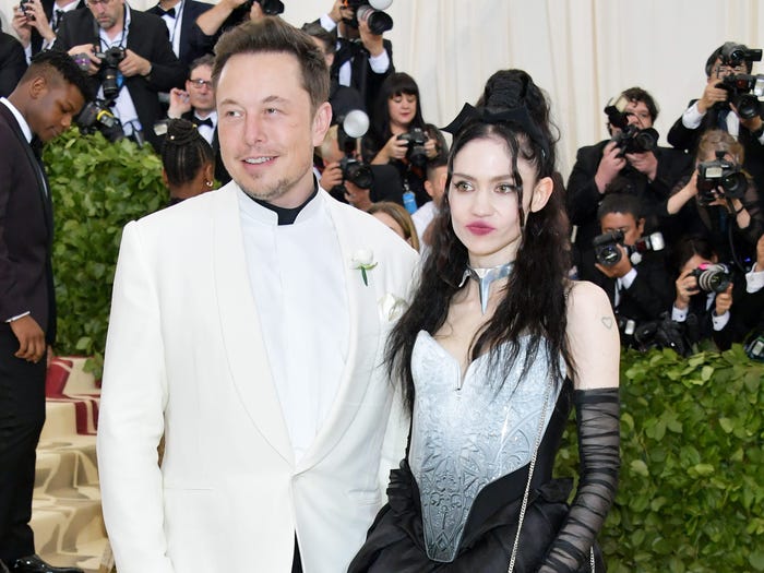 Elon Musk and Grimes standing together at the 2018 Met Gala. Musk is wearing a white blazer and Grimes is wearing a long dress with a black skirt.