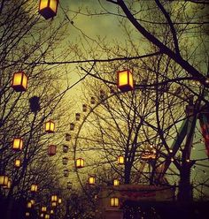 lanterns are hanging from the branches of trees in front of a ferris wheel at night