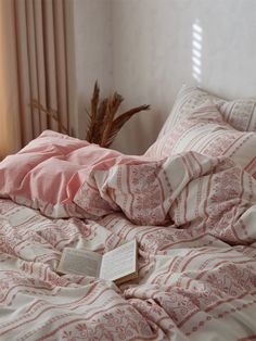 an open book sitting on top of a bed covered in pink and white sheets next to a window