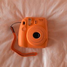 an orange polaroid camera sitting on top of a white bed with a red cord