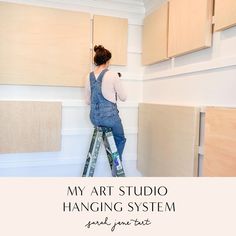 a woman on a ladder painting the walls in her kitchen with text overlay that reads, my art studio hanging system paint your first