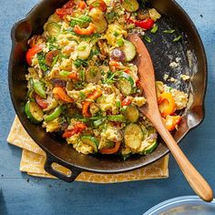 a skillet filled with vegetables on top of a blue counter next to a wooden spoon