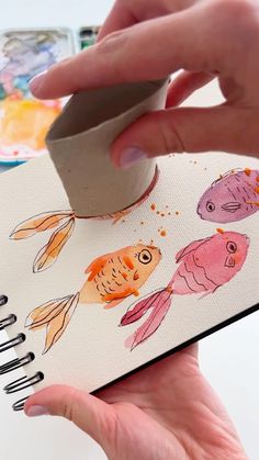 a hand is holding an open notebook with watercolor fish on it and another drawing in the background