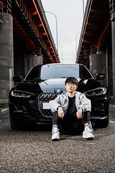 a young man sitting on the ground next to a black bmw parked under a bridge