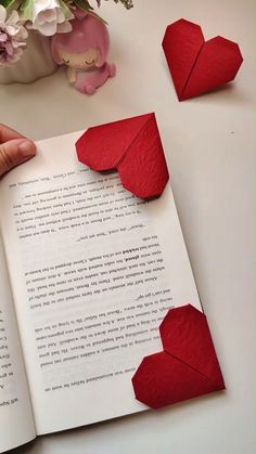 an open book with red paper hearts on the page and flowers in vase next to it