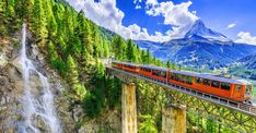 a red train traveling over a bridge next to a waterfall in the middle of mountains