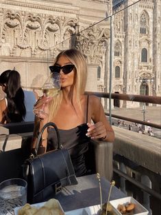 a woman sitting at an outdoor table drinking from a wine glass in front of a building