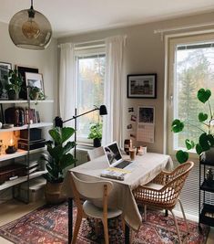 a living room filled with furniture and plants next to two large windows on top of a wooden floor