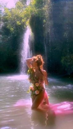 a woman in a pink dress standing in the water near a waterfall with flowers on it