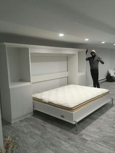 a man standing next to a bed in a room with white walls and wooden floors