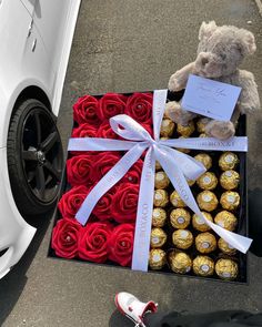 a teddy bear sitting on top of a box filled with roses and chocolates next to a white car