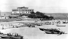 black and white photograph of people on the beach with boats in front of a large building