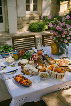 a table with food and wine on it in front of a house, next to some flowers