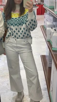 a woman taking a selfie with her cell phone in a store aisle, she is wearing white pants and a patterned sweater