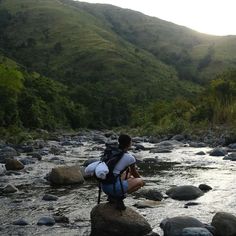 a person sitting on top of a rock in the middle of a river surrounded by mountains