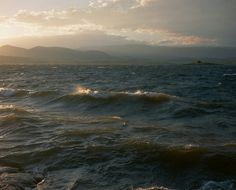 the sun is setting over the ocean with mountains in the backgrouund and clouds in the sky