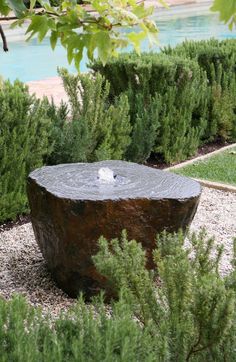 an outdoor fountain in the middle of a garden with trees and shrubs around it, near a pool