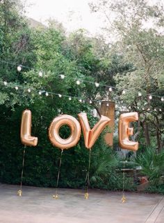 some balloons that say love are hanging in front of trees and bushes with string lights