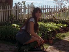 a woman sitting on the ground next to a tree and potted plants in front of a white picket fence