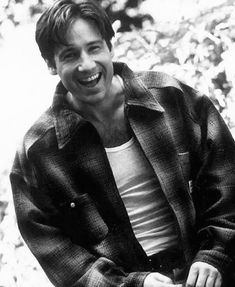 black and white photograph of a man sitting on a bench smiling at the camera with his hands in his pockets