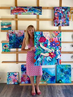 a woman standing in front of some colorful paintings on a wooden wall with she is holding up the painting