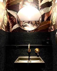 a bathroom sink under a round mirror in front of a wallpapered wall with leaves on it