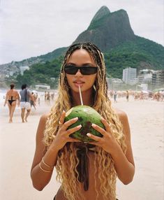 a woman drinking a drink while holding a green coconut in front of her face on the beach