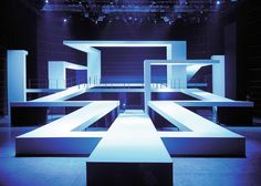 an empty stage with blue lighting and white benches in the center, on which is a chandelier