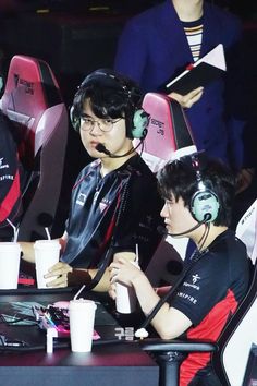 two young men sitting at a table with headphones on and one holding a drink