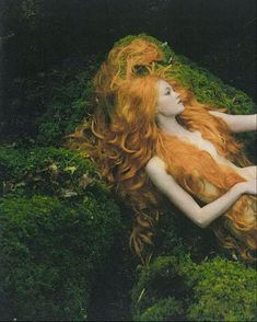 a woman with long red hair laying on top of green plants in the grass,