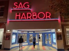 two people are walking in front of the sag harbor store at night, with neon lights