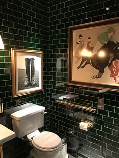 a bathroom with green tiled walls and black tile flooring, two framed pictures above the toilet