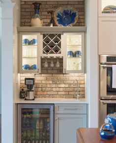 a kitchen with a brick wall and white cupboards filled with wine bottles, glasses and other items