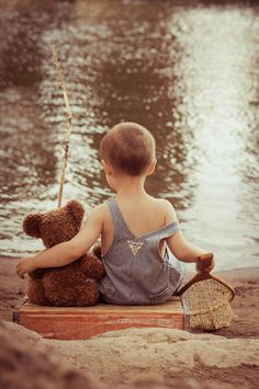 a little boy sitting in front of the water with a teddy bear and tennis racket