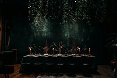 a long table with candles on it in front of a dark wall and plants hanging from the ceiling